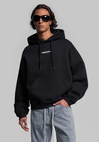 Felpa con cappuccio nera, con tasca frontale e logo bianco. Modello che indossa occhiali da sole scuri e jeans chiari strappati. Texture liscia.