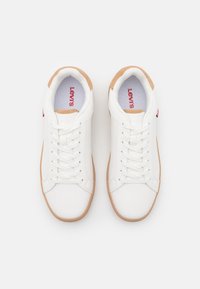 Białe trampki z gładką skórzaną powierzchnią, kontrastującą brązową gumową podeszwą oraz czerwonym logo "Levi's" na języku i boku. Design sznurowany.
