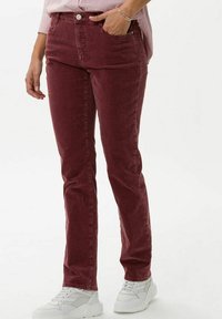 Femme portant un pantalon en velours côtelé bordeaux taille haute et jambes droites, associé à des baskets blanches et une chemise rose rayée.