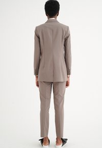 Abito color taupe chiaro con giacca sartoriale e pantaloni attillati. Tessuto morbido, rever a nocciola e dettagli con tre bottoni sulle maniche.