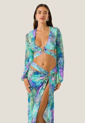 Donna che indossa un vibrante abito multicolore con ritagli, maniche lunghe, dettagli ad anello e uno spacco alto che mostra la sua gamba su uno sfondo semplice.