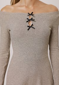 Off-shoulder ribbad klänning i ljusgrått med tre svarta rundade rosetter vid halsringningen och långa åtsittande ärmar. Texturerad tyg.