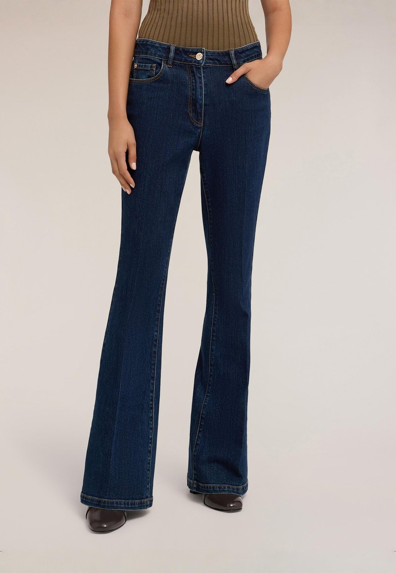 Žena nosící tmavě modré džíny s vysokým pasem a rozšířenými nohavicemi, jedna ruka v kapse, v hnědých botách, stojící před jednobarevným pozadím.