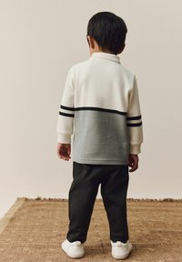 Langes Oberteil mit weißen und grauen Streifen und strukturiertem Stoff, kombiniert mit schwarzen Hose und weißen Sneakers, von hinten betrachtet.