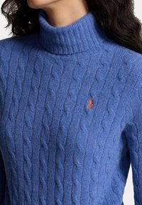 Sininen palmikkoneuleinen pooloneule, jossa on resorikaulus ja oranssi hienovarainen logodetaili. Pehmeä rakenne ja monimutkaiset neuleet luovat kodikkaan ilmeen.