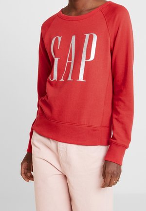 Rød langærmet sweatshirt med store sølvfarvede "GAP"-bogstaver på forsiden, båret med lyserøde bukser på en stående person.