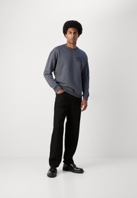 Sivi dukserica s okruglim izrezom i logom na prsima, u kombinaciji s crnim hlačama ravnog kroja i crnim cipelama na jednobojnoj pozadini.