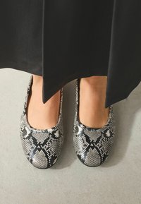 Ploché topánky s čierno-bielym hadím vzorom, zaoblenou špičkou a hladkou textúrou, umiestnené na neutrálnom povrchu pod čiernym odevom.