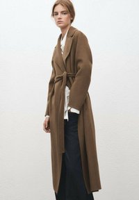 Długi brązowy płaszcz z wełny z paskiem w talii, klapami z wycięciem oraz pojedynczym zapięciem na guzik, noszony na białej koszuli i ciemnych dżinsach.