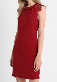 Robe rouge sans manches ajustée, longueur genou, avec des manches en dentelle, portée par une femme debout devant un fond blanc.