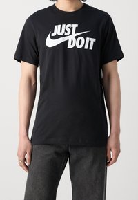 En person som bär en svart T-shirt med vit text "JUST DO IT" och Nike-swoosh-logotyp, kombinerad med mörkgrå jeans mot en neutral bakgrund.