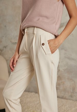 Beżowe spodnie o dopasowanym kroju z gładką fakturą, z bocznymi kieszeniami z guzikowymi klapami i zamkiem błyskawicznym. Połączone z różowym swetrem.