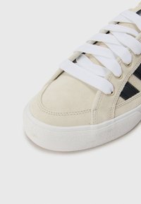 Beige semsket sneakers med hvite snørebånd, en hvit gummisåle, og svarte diagonale striper som fremhever det øvre designet.
