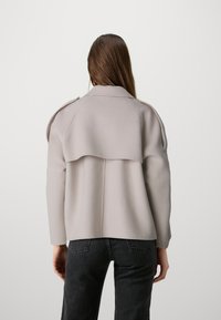 Giacca in misto lana grigio chiaro con design a pattina sulle spalle, silhouette strutturata e maniche lunghe, abbinata a jeans scuri.