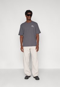 Camiseta cinza oversized de manga curta com um acabamento texturizado e um logótipo branco impresso no lado esquerdo do peito, combinado com calças largas de cor creme.