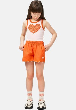 Biały top na ramiączkach z pomarańczową grafiką serca, w połączeniu z pomarańczowymi szortami z falistym dołem i haftowanym detalem; noszone z podkolanówkami.