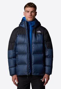 Granatowa kurtka puchowa z czarnymi ramionami, kapturem i suwakami. Posiada pikowany wzór, dwie boczne kieszenie oraz logo na klatce piersiowej.