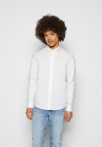 Camicia bianca a maniche lunghe con bottoni, dal tessuto morbido, design aderente e orlo curvo, abbinata a jeans azzurri chiari.