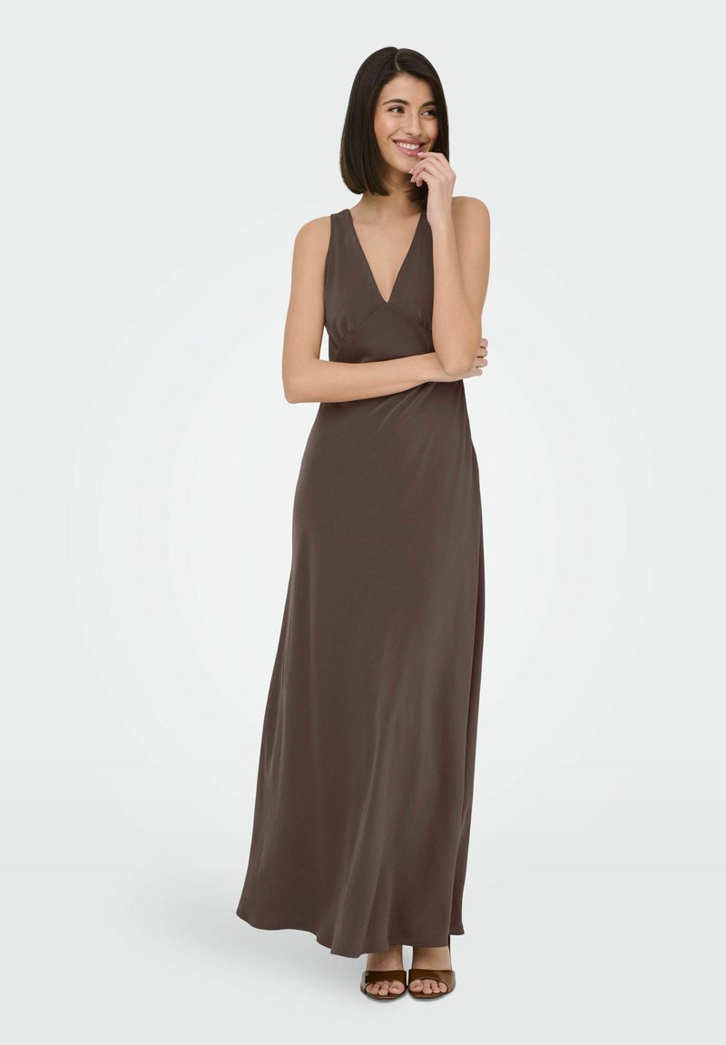 Femme portant une robe longue marron sans manches avec un décolleté en V, souriante, debout, une main touchant son menton, l'autre croisée sur son corps.