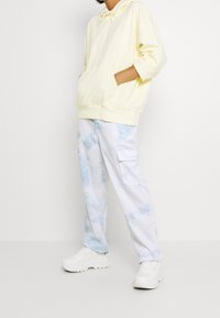 Felpa gialla con cappuccio e tasca frontale, abbinata a pantaloni cargo azzurri tie-dye e sneakers bianche. Look casual e vestibilità rilassata.