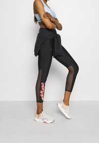 Černé legíny s krajkovými panely a růžovým grafickým potiskem, spárované se světle modrým sportovním podprsenkou a bílými vzorovanými sportovními botami.