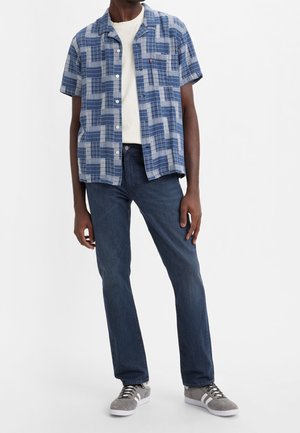 Homme portant une chemise à manches courtes bleue à motifs par-dessus un t-shirt blanc, un jean bleu foncé et des baskets grises avec des semelles blanches.