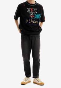 Persona con camiseta gráfica negra con diseño abstracto colorido, jeans oscuros y zapatillas beige, de pie con las manos en los bolsillos.