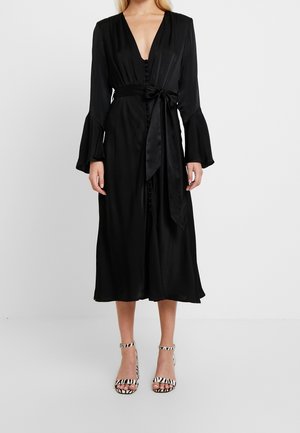 Zwarte zijden jurk met een diepe V-hals, lange klokmouwen en een gestrikt taille. Voorzien van knopen aan de voorkant en een midi-lengte zoom. Een paar gestreepte sandalen.