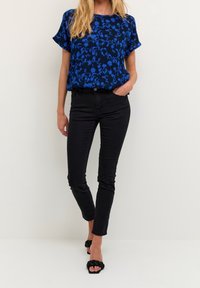 Blå bluse med blomstermønster og korte ærmer, parret med sorte stramme jeans og sorte flettede slidesandaler. Glat stofstruktur.