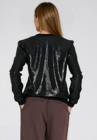 Czarna kurtka z cekinami o fakturowanej powierzchni, charakteryzująca się luźnym krojem, ściągaczami na mankietach i dole oraz gładką podszewką. Widok z tyłu ukazuje cekiny na całej powierzchni.