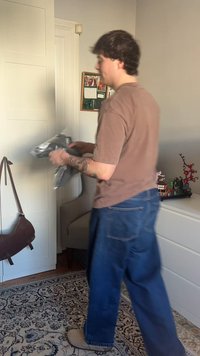 Personne portant une chemise marron et un jean bleu tenant des vêtements sombres pliés dans une pièce avec des meubles blancs et un tapis décoratif.