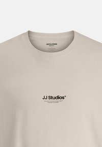 Lys beige bomulds t-shirt med rund hals og et trykt logo i sort på brystet. Enkel, afslappet pasform med glat tekstur.