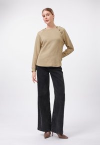Felpa beige a fantasia con tessuto testurizzato e bottoni decorativi, abbinata a jeans neri a vita alta e gamba larga, e tacchi a punta color sabbia.