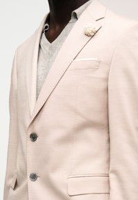 Veste de costume beige à deux boutons, un pull à col en V gris clair en dessous, un mouchoir de poche blanc et une broche fleur ivoire sur le revers.