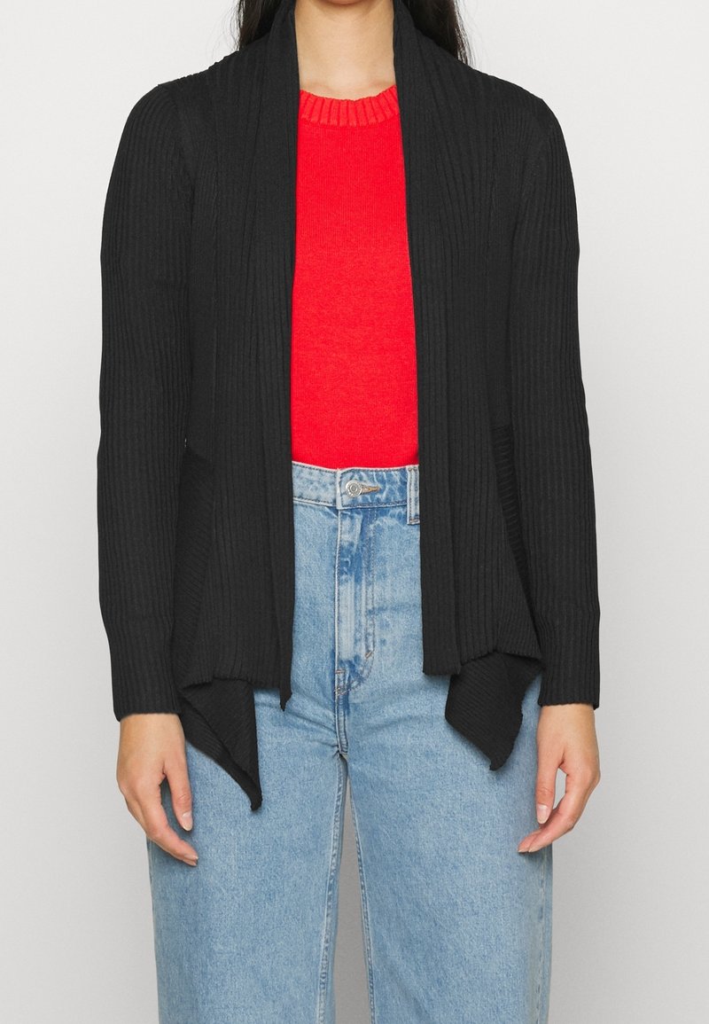 Femme portant un cardigan noir à côtes ouvert sur un pull rouge et un jean bleu clair taille haute, debout les bras détendus.