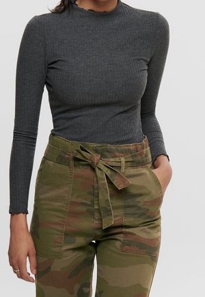 Femme portant un haut moulant à manches longues gris foncé et un pantalon taille haute camouflage avec une ceinture en tissu nouée, main dans la poche.