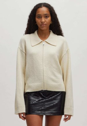 Crème-kleurige zip-up sweater met een grote kraag, lange mouwen, geribde manchetten en een zachte textuur. Gemaakt over een zwarte neplederen rok.