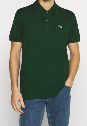 Homme portant un polo à manches courtes vert foncé avec deux boutons et un petit logo crocodile sur la poitrine, associé à un jean bleu.