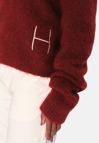 Rød, luftig genser med ribbede mansjetter, med en hvit brodert "H" foran. Teksturen ser myk og koselig ut.