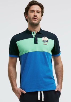 Mann trägt ein kurzärmeliges Polo mit schwarzen, mintgrünen und blauen horizontalen Farbblöcken, steht mit den Händen in den Taschen.