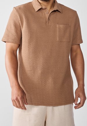 Mann trägt ein kurzärmliges, strukturiertes braunes Poloshirt mit Tasche auf der linken Brust und hellen Hosen.