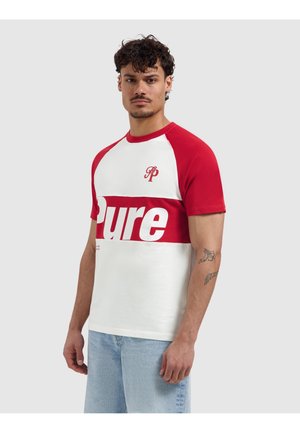 Man met krullend haar, gekleed in een wit-rood T-shirt met korte mouwen met de tekst "Pure" en lichtblauwe spijkerbroek, staand tegen een effen achtergrond.