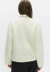 Sweter w kolorze kremowym o miękkiej, puszystej teksturze, z długimi rękawami i kołnierzem. Hem jest lekko zaokrąglony, pokazany od tyłu.