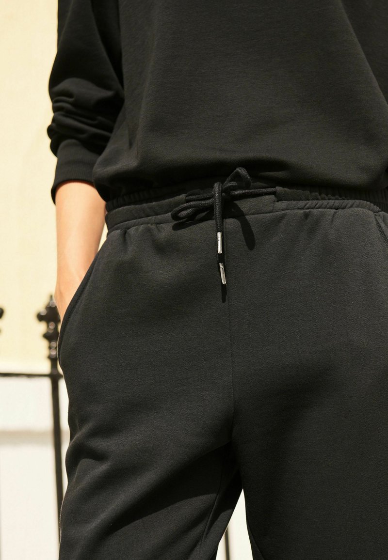 Schwarze Sweatpants mit elastischem Bund, Kordelzug und Seitentaschen. Aus weichem Baumwollmischgewebe mit glatter Textur gefertigt.