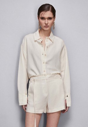 Camicetta leggera a maniche lunghe color crema con bottone frontale e polsini. Abbinata a pantaloni corti a vita alta coordinati con cordoncino.