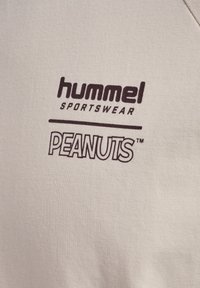 Beige katoenen T-shirt met donkerbruine bedrukte logo's van "hummel SPORTSWEAR" en "PEANUTS" in blok- en gestileerde tekst.