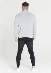 Pull en tricot gris à manches longues, associé à un jean slim noir et des baskets blanches, vu de dos. Tatouage de cou notable visible.