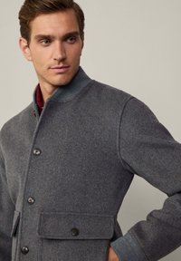 Chaqueta de mezcla de lana gris con cuello alzado, cierre frontal de botones, bolsillos laterales y puños acanalados. La textura parece suave y cálida.