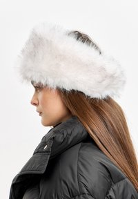 Bijela lažna krzna traka za glavu s pahuljastom teksturom, nosi se preko dugih smešnih kose, u kombinaciji s crnom jaknom od puffera. Pogled sa bočne strane.