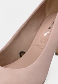 Κοντινό πλάνο ενός ψηλού τακουνιού από ελαφρύ ροζ σουέντ που δείχνει την ομαλή εσωτερική επένδυση και μέρος της εσωτερικής σόλας με το κείμενο της μάρκας.